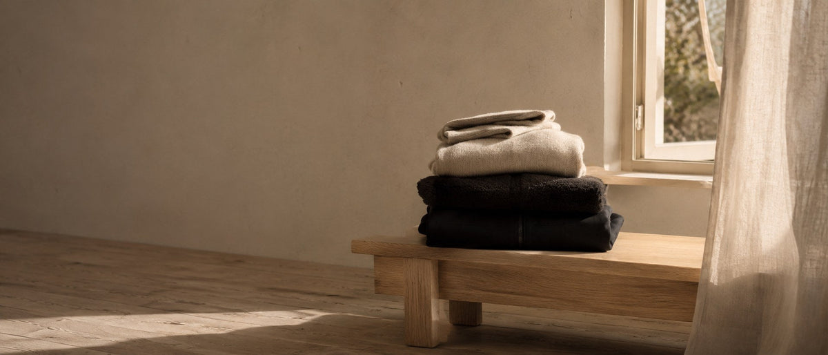 Folded knitwear and outerwear stacked on a pale oak bench beside an open window