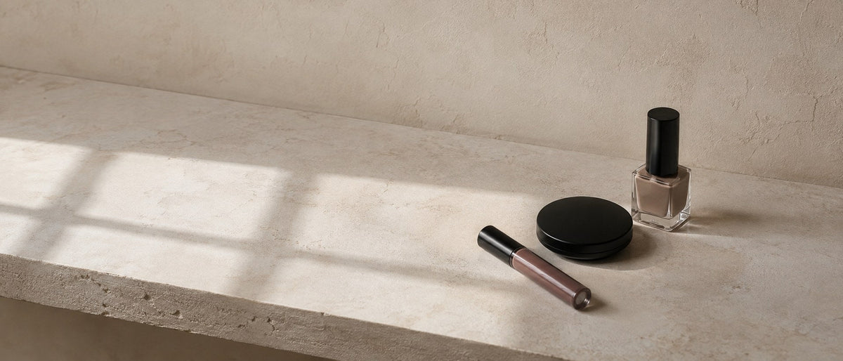 An unbranded lip gloss, black compact and bottle of nude nail polish on a travertine ledge in raking window light