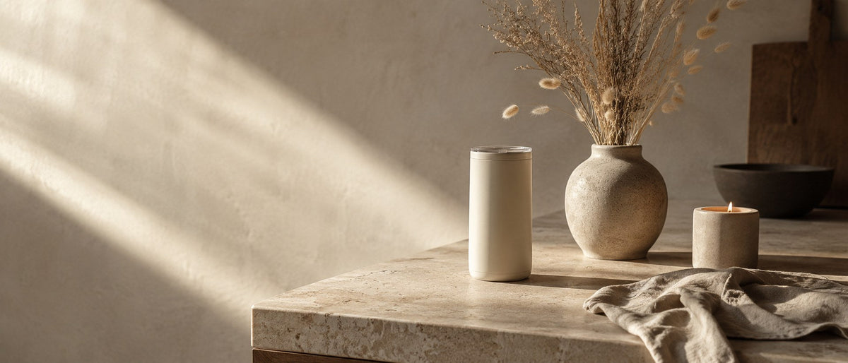 An insulated tumbler, ceramic vase of dried grasses and a lit candle on a travertine counter in morning light