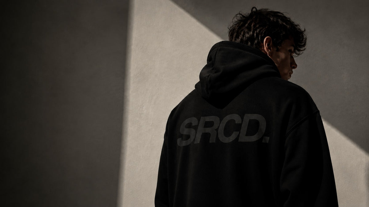 Model seen from behind wearing a black SRCD oversized hoodie against a sunlit concrete wall
